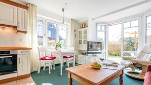 A bright kitchen with white cabinets, a dining area with a table and chairs, a TV, and a large window with a view of the garden. - Komfortabel. Strandnah. Zentral. Objekt-id: Has/09 (Westerland)
