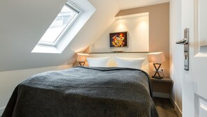 A bedroom with a skylight, a bed with a dark blanket, two bedside tables with lamps, and a colorful painting on the wall. - Sauna. Exquisit. Stimmig (Westerland)