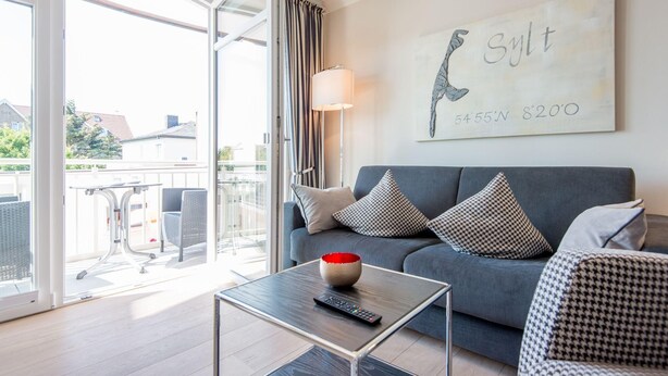 A modern living room with a grey sofa, patterned cushions, a glass coffee table with a remote control, and a wall-mounted artwork. - Modern. Lichtdurchflutet. Wellness (Westerland)