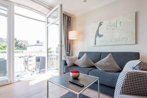 A modern living room with a grey sofa, patterned cushions, a glass coffee table with a remote control, and a wall-mounted artwork. - Modern. Lichtdurchflutet. Wellness (Westerland)