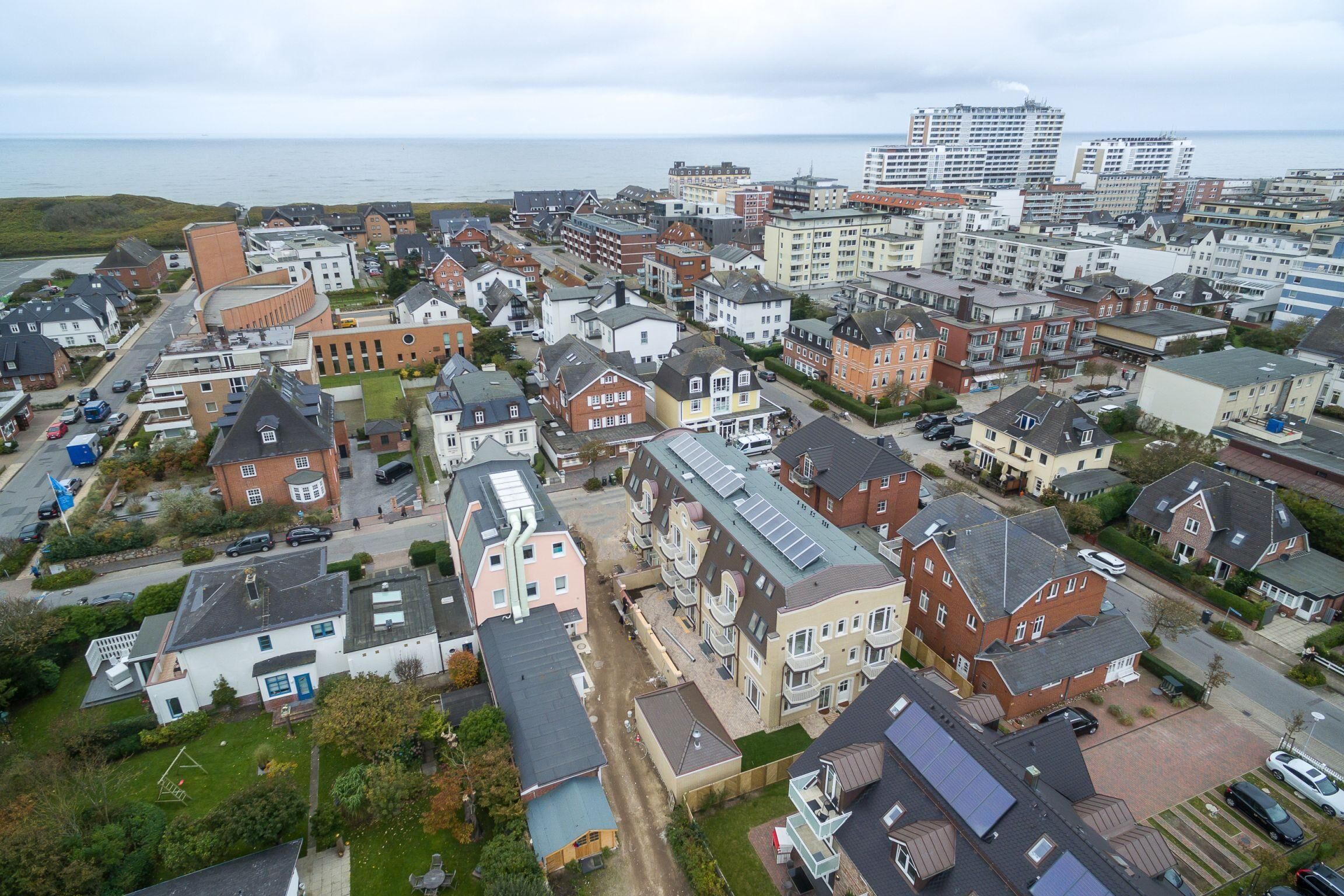 An aerial view of a coastal residential area with various buildings, including houses and apartment complexes, with a view of the sea in the background.
