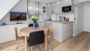 Une cuisine moderne avec des meubles blancs, une table à manger en bois accompagnée de quatre chaises, une grande fenêtre avec des rideaux et un téléviseur fixé au mur.