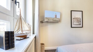 Ein Zimmer mit einem Bett, einem Vorhangfenster, einem an der Wand befestigten Fernseher sowie einem kleinen Tisch mit Büchern und einem Modellsegelboot.