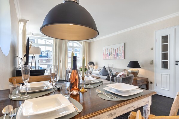 Un comedor con una mesa de madera dispuesta para cuatro, una lámpara colgante negra grande y vista al salón en el fondo.