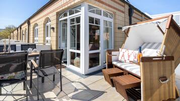 A sunlit patio with a seating area, a glass-enclosed room, and a brick building facade.