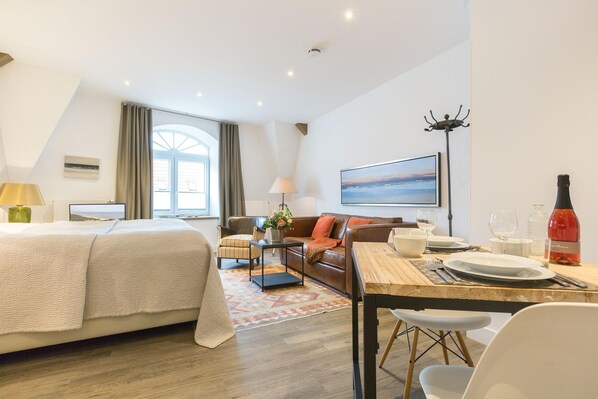 Une chambre d’hôtel moderne avec un grand lit, un canapé, une table à manger pour deux personnes et une grande fenêtre avec des rideaux.