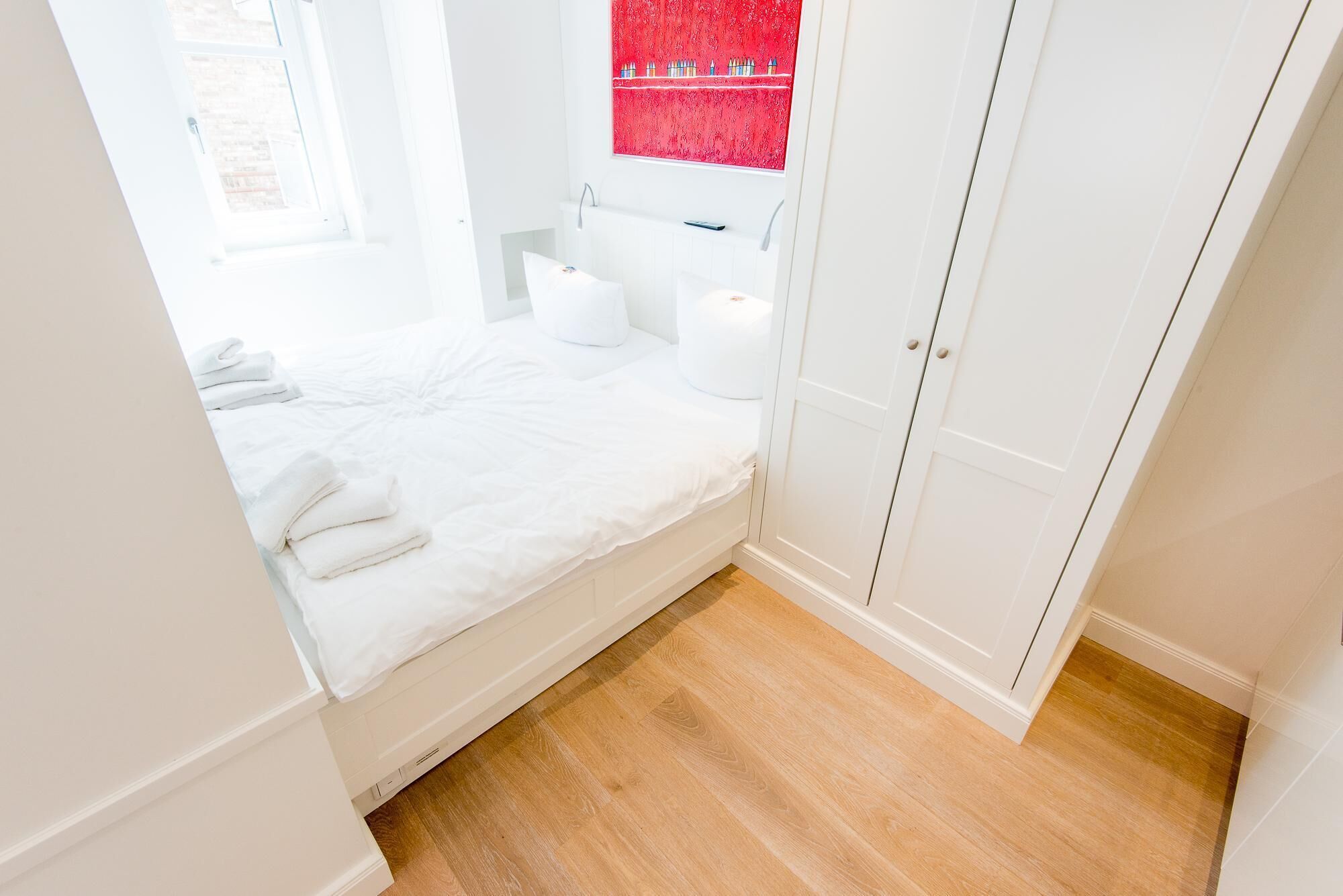 A small, bright bedroom with a single bed, a white wardrobe, and a window with a red view.