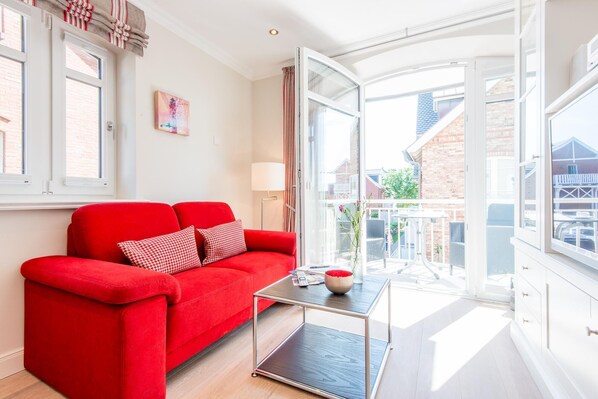 Une pièce lumineuse avec un canapé rouge, une petite table et une grande fenêtre donnant sur un balcon et les bâtiments environnants.