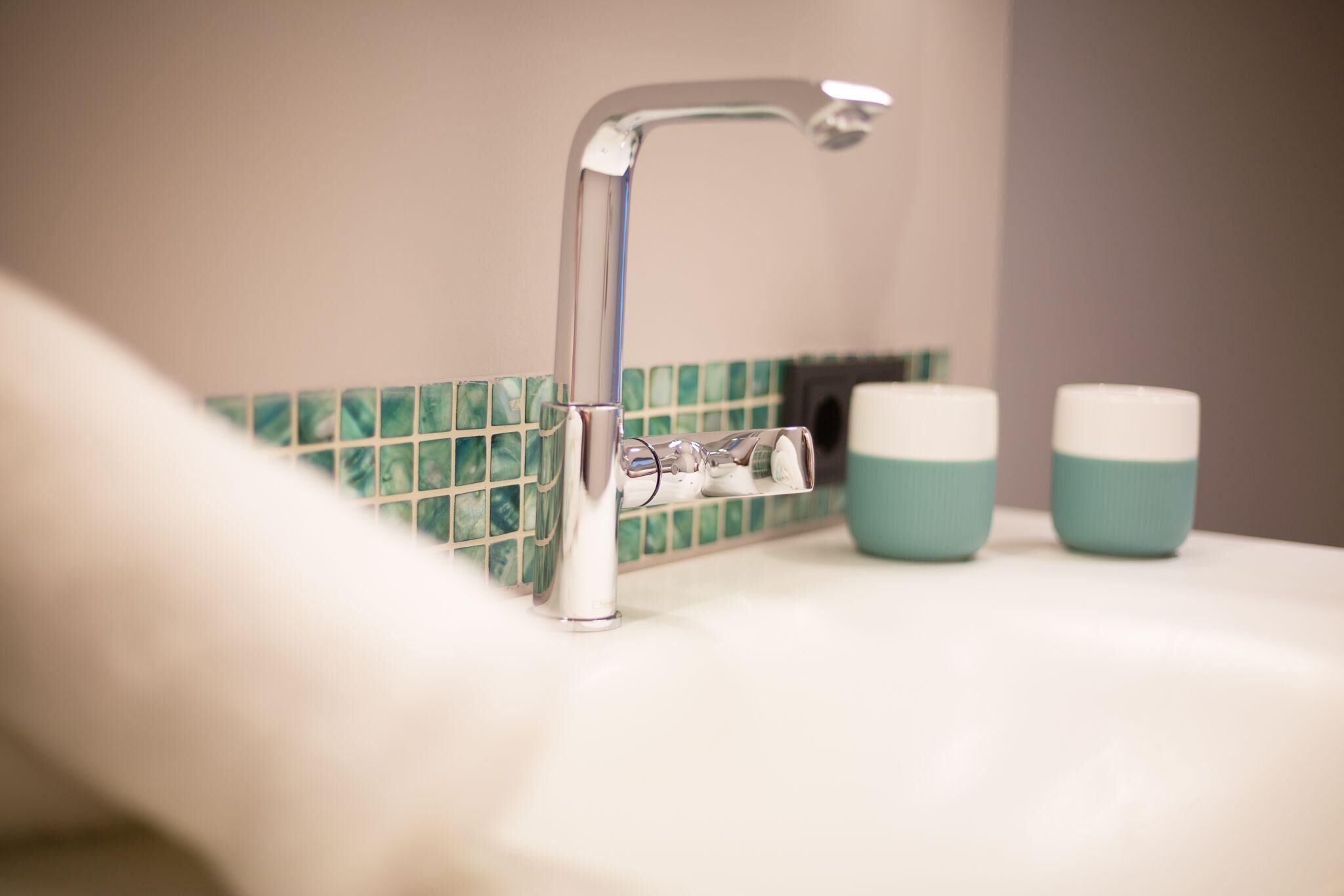 A bathroom sink with a chrome faucet, a tiled backsplash, and two teal and white cups on the counter.