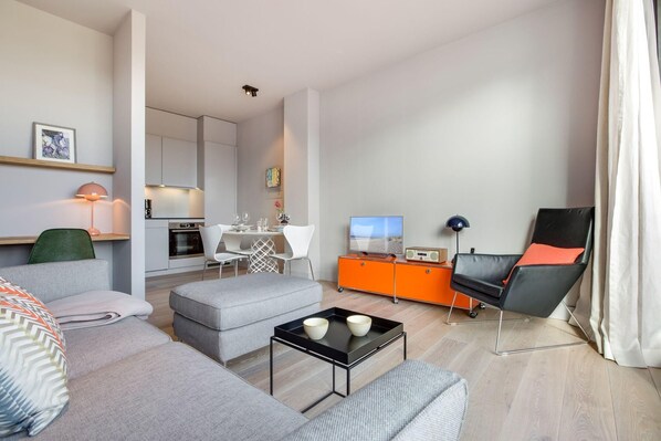 Un salon moderne avec un canapé, un pouf et une table basse. On aperçoit un téléviseur, un fauteuil et un meuble en arrière-plan.
