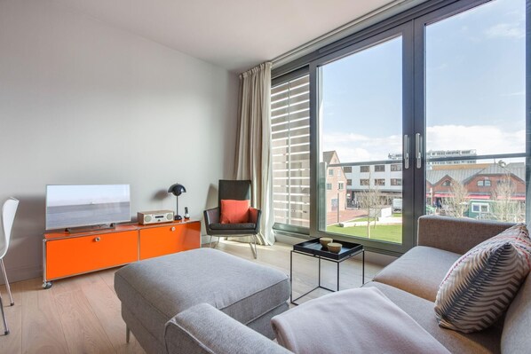Una habitación de hotel moderna con un ventanal grande, un televisor de pantalla plana, un minibar naranja, un sofá gris y una silla negra con un cojín rojo.