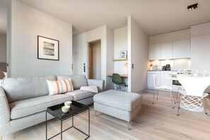 Un salon moderne avec un canapé gris, une petite table et un coin cuisine doté de meubles blancs et d’une table à manger.