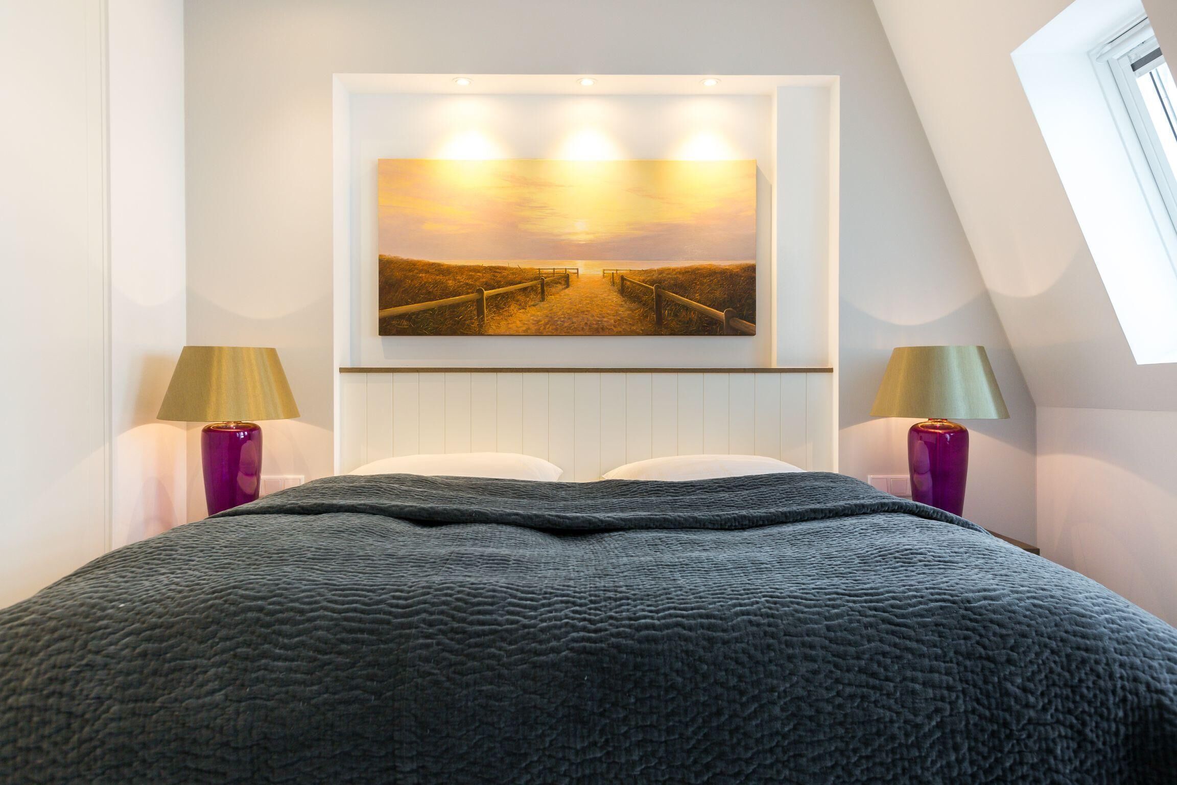 Ein Schlafzimmer mit einem großen Bett, einem Gemälde von einem Sonnenuntergang über einem Strand und je einer Lampe auf jeder Seite.