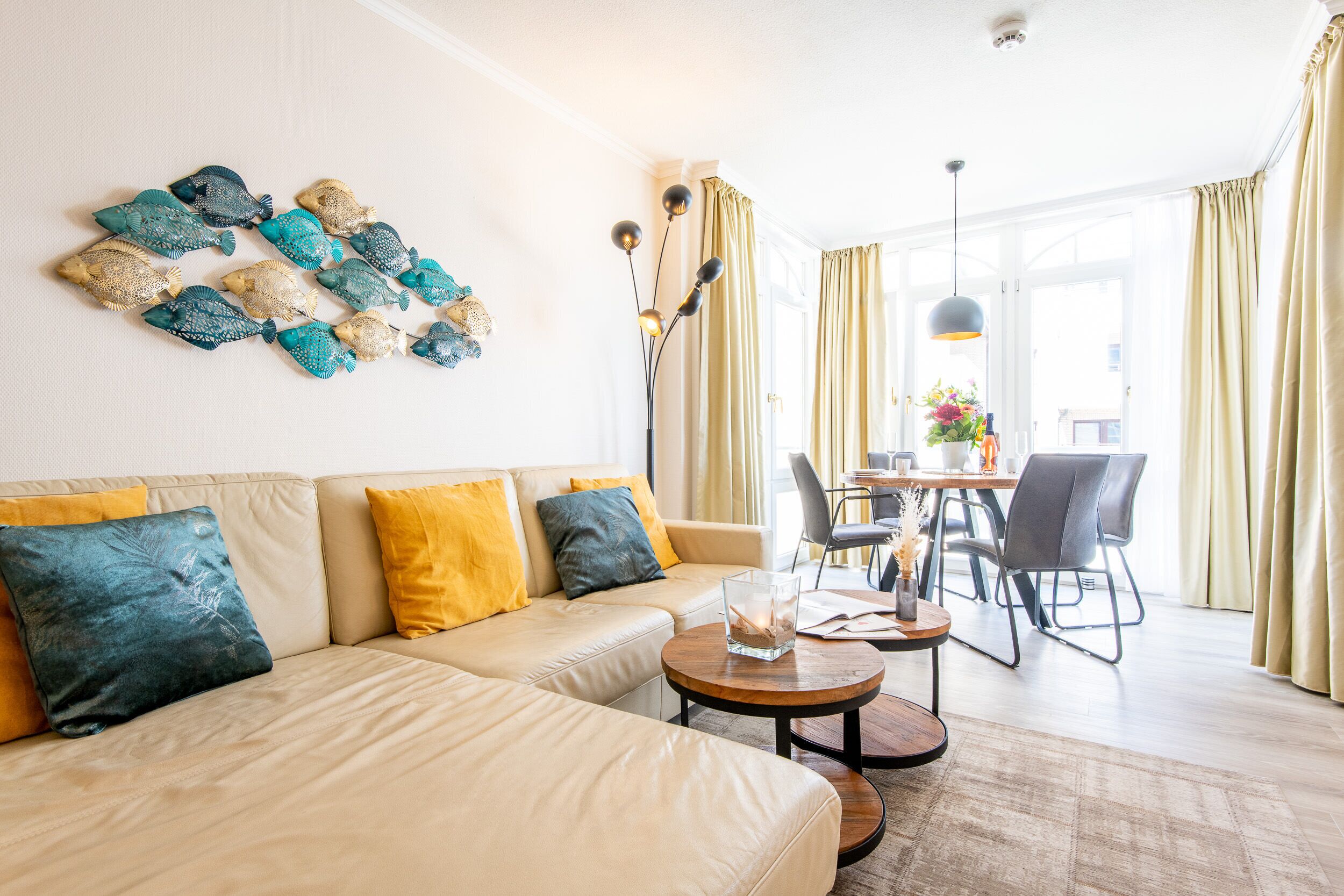 Ett modernt vardagsrum med en beige soffa, gula och blå kuddar, ett soffbord och en matplats med stolar och ett bord.