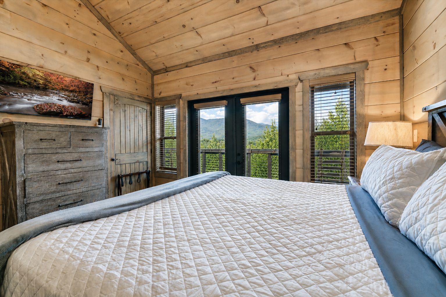 A bedroom with a large bed, wooden walls, a dresser, and a window with a view of greenery.