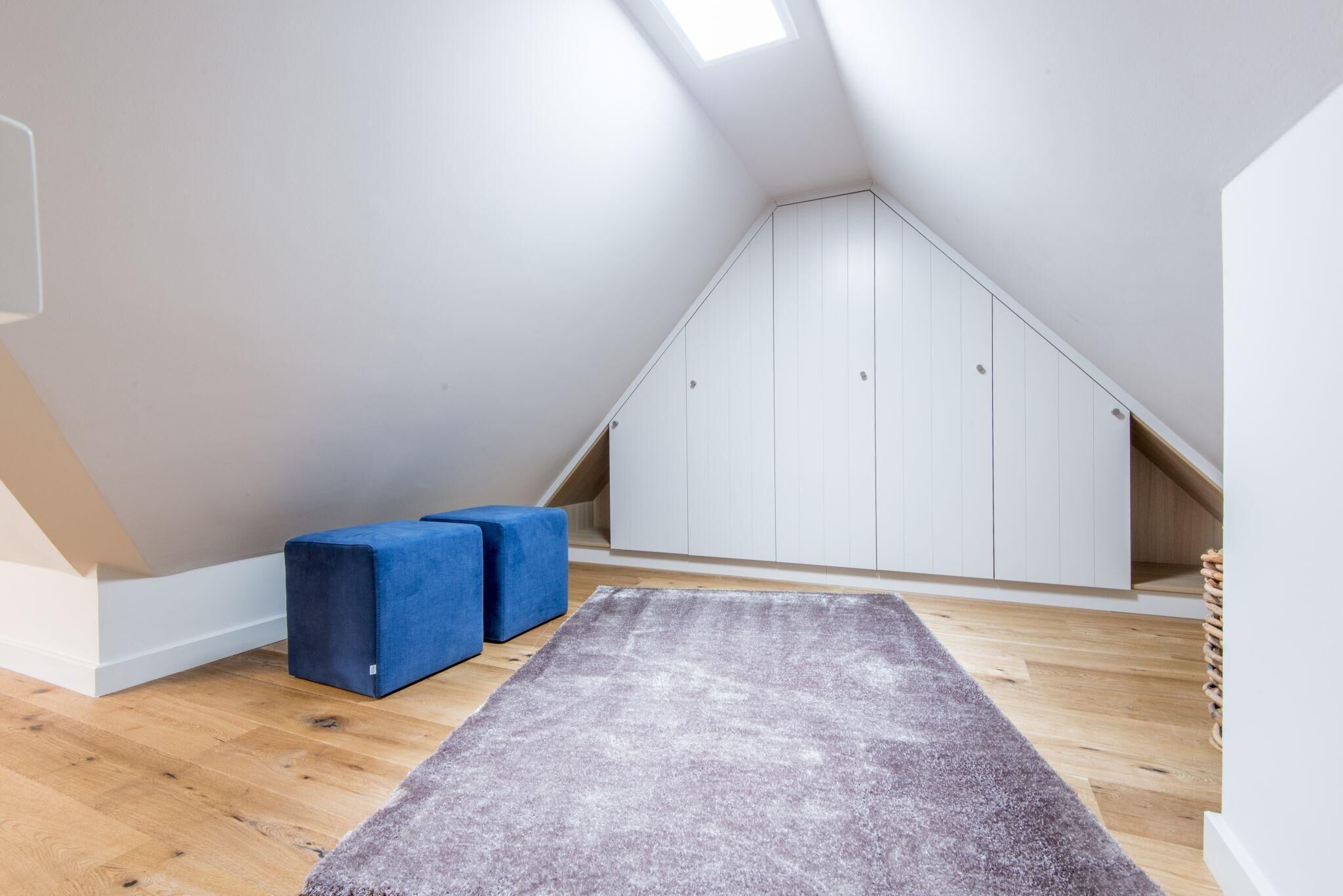 A room with a sloped ceiling, wooden flooring, a grey carpet, and a white wardrobe.
