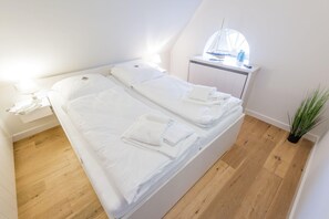 A neatly made bed with white linens and towels, a bedside table with a lamp, a small cabinet, and a window with a sailboat model.