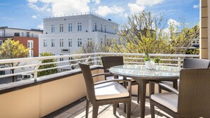 Un balcon doté de meubles en osier et d’une table en verre, offrant une vue sur un immeuble et des arbres.