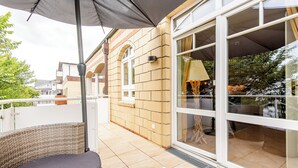 Ein Balkon mit einem Korbstuhl, einem Tisch mit Zeitschriften und einem großen Fenster mit Blick auf den Garten.