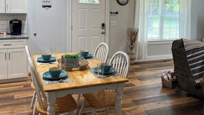 Uma sala de jantar com mesa de madeira posta para quatro, cadeiras brancas, área de cozinha com armários brancos e janela com cortinas brancas.