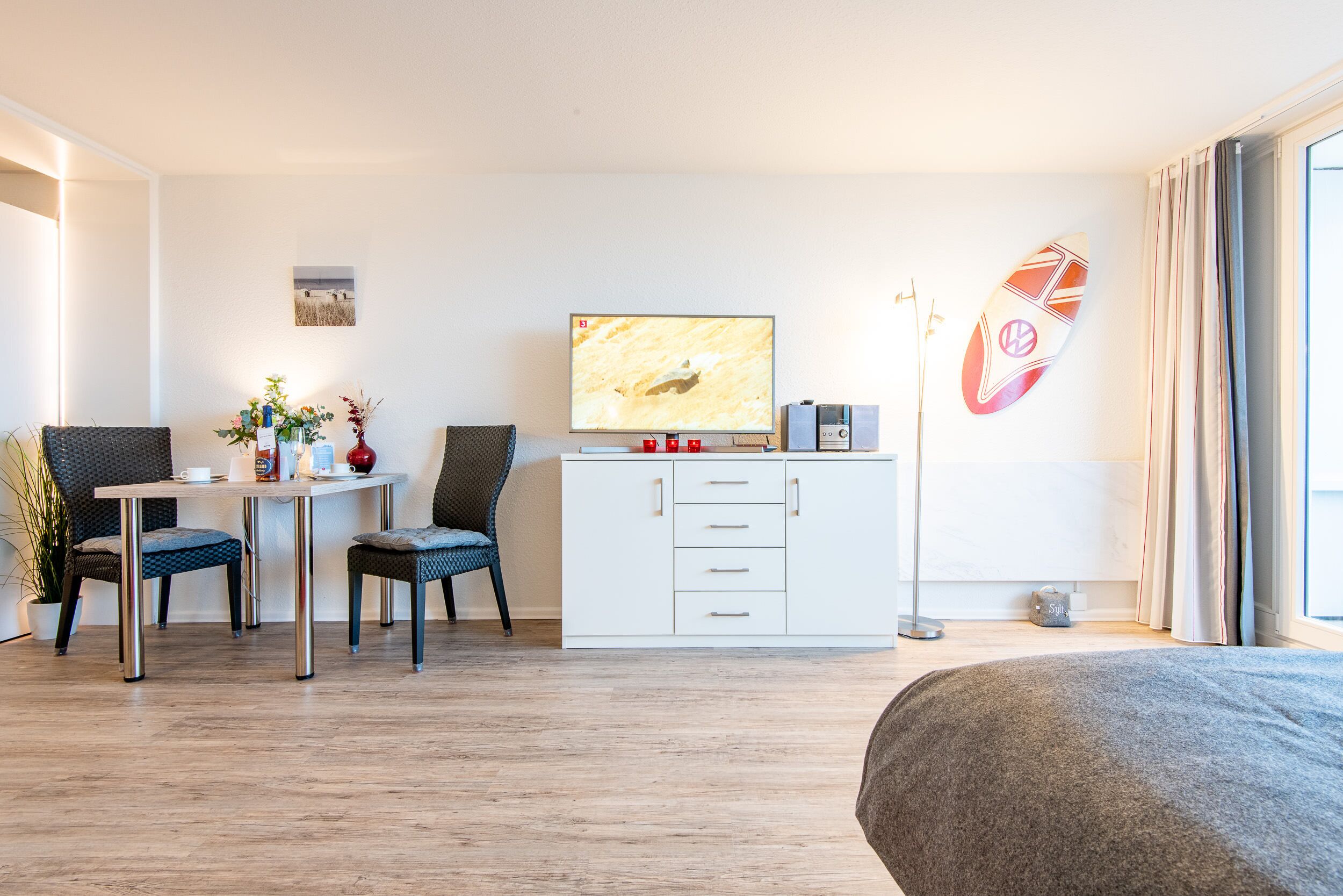 A modern hotel room with a wooden floor, a bed, a desk with two chairs, a television, a white cabinet, and a surfboard decoration.