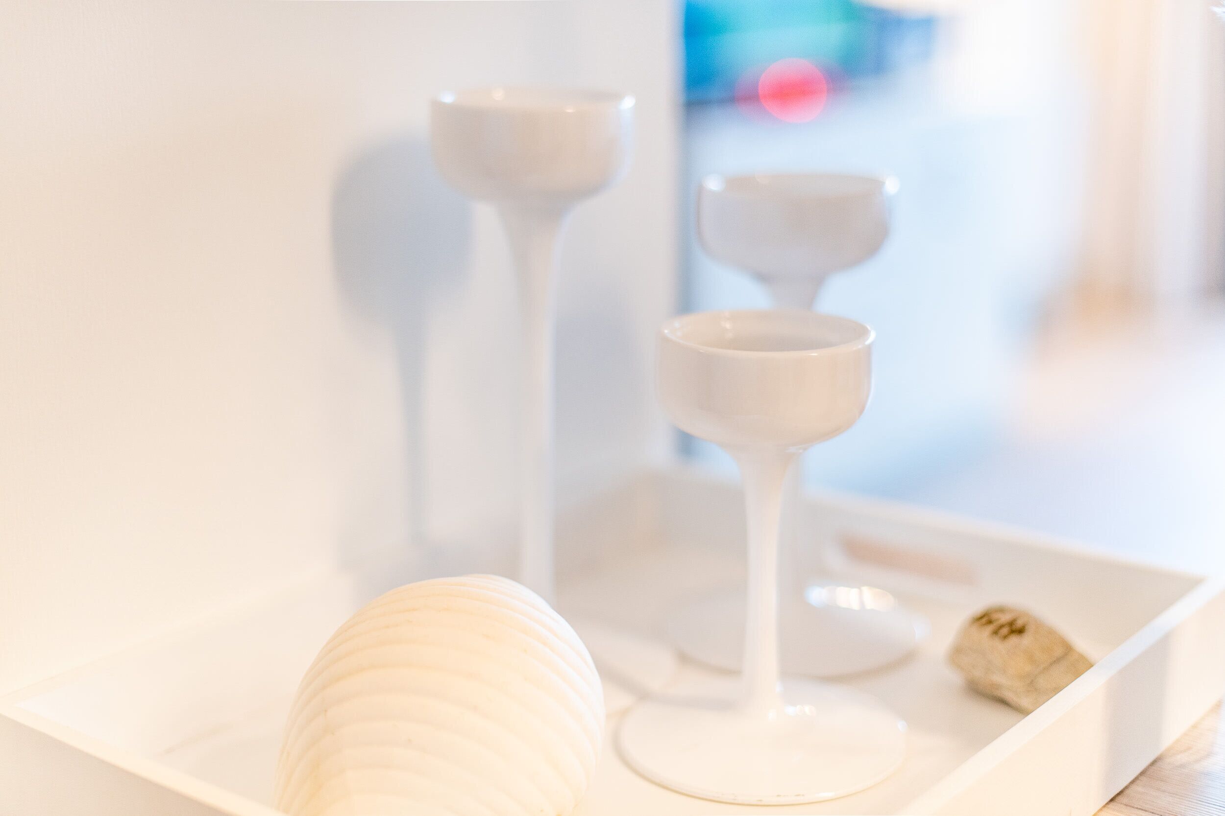 Un plateau blanc avec une bougie, deux verres à vin blanc et un petit objet sur une surface blanche.