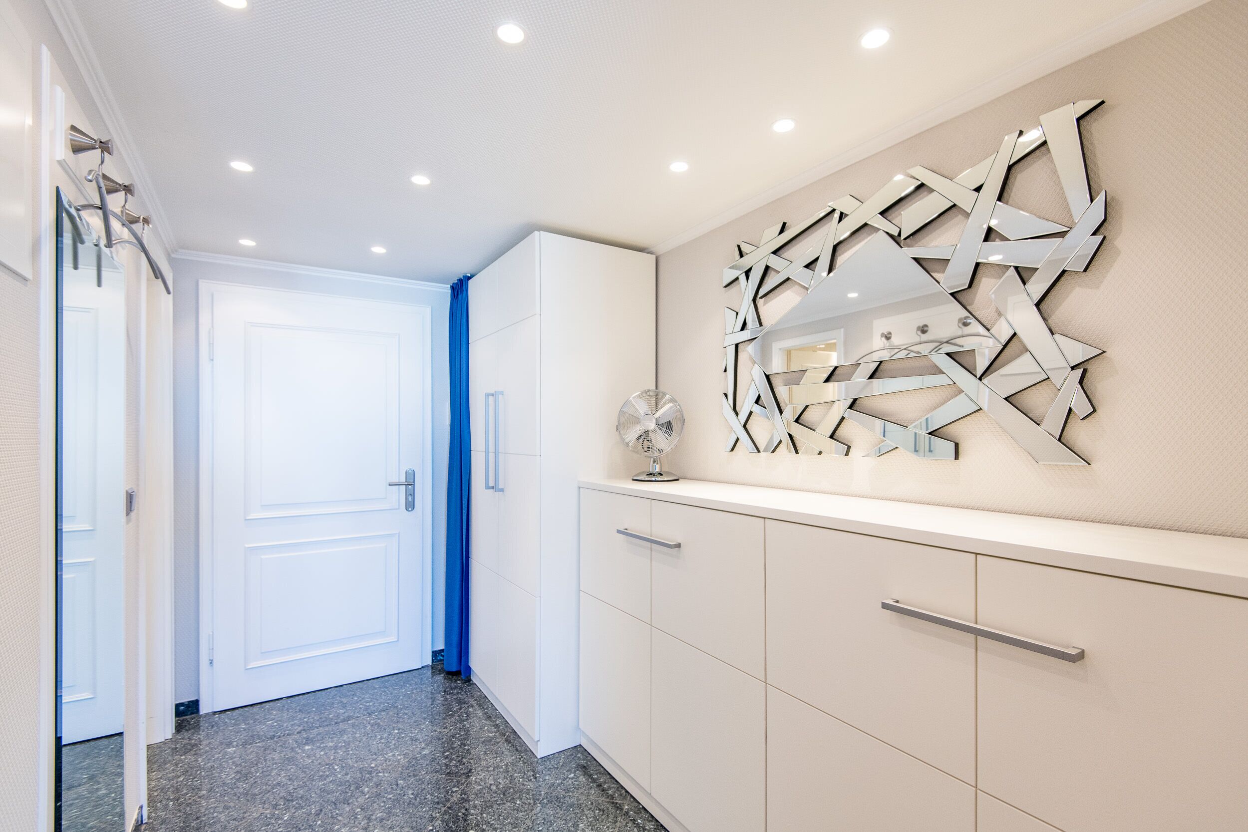Um corredor com porta branca, cortina azul, ventoinha na parede e um espelho geométrico decorativo.