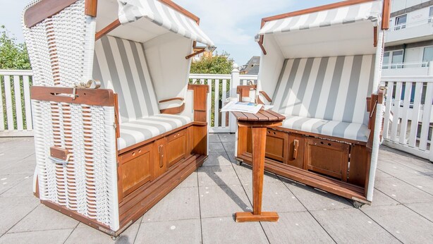 Dos sillones de madera con cojines a rayas y toldos blancos, ubicados en un patio con baldosas y una valla blanca al fondo.