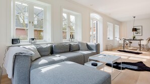 A modern living room with a grey sectional sofa, a white coffee table, and a black fur rug. There are large windows providing natural light and a view of the outside. - Dansk Hus Sylt List (List)