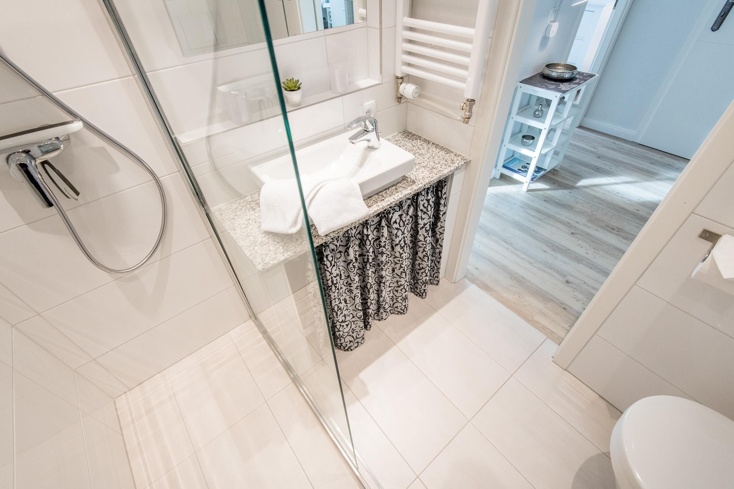 Un baño moderno con cabina de ducha de vidrio, un lavamanos blanco y un mueble de tocador con cortinas estampadas.