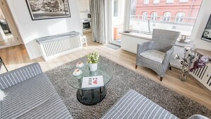 Ein modernes Wohnzimmer mit einem Glastisch, gestreiften Sofas, einer Topfpflanze und Blick auf ein rotes Backsteingebäude durch ein großes Fenster.