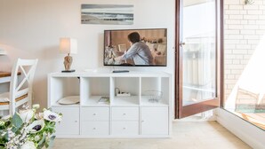Una sala moderna con un mueble blanco para el equipo multimedia, un televisor de pantalla plana, una lámpara y un ventanal con vista a la playa.