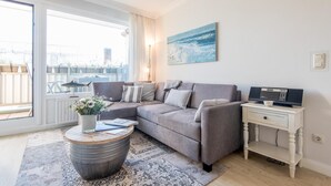 Una sala de estar moderna con un sofá seccional gris, una mesa de centro redonda con un jarrón, una mesita de noche blanca con una radio y un ventanal con vistas a edificios y zonas verdes.
