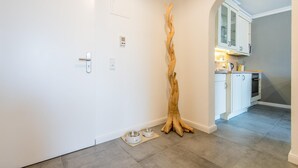 Een gang met een witte deur, een houten sculptuur en een kijkje in een keuken met witte kasten en een betegelde vloer.