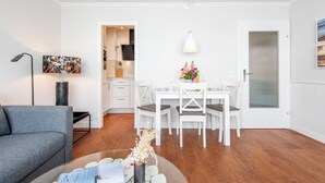 A modern living room with a grey sofa, a dining table with four chairs, a white wall, and a kitchen in the background. - Gemütlich. Zentral. Strandnah. Objekt-id: Hdb/24 (Westerland)