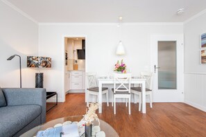 Un salon moderne avec un canapé gris, une table à manger avec quatre chaises, un mur blanc et une cuisine en arrière-plan.