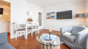 A modern living room with a glass coffee table, a grey sofa, a dining area with white chairs, and a flat-screen TV mounted on the wall. - Gemütlich. Zentral. Strandnah. Objekt-id: Hdb/24 (Westerland)