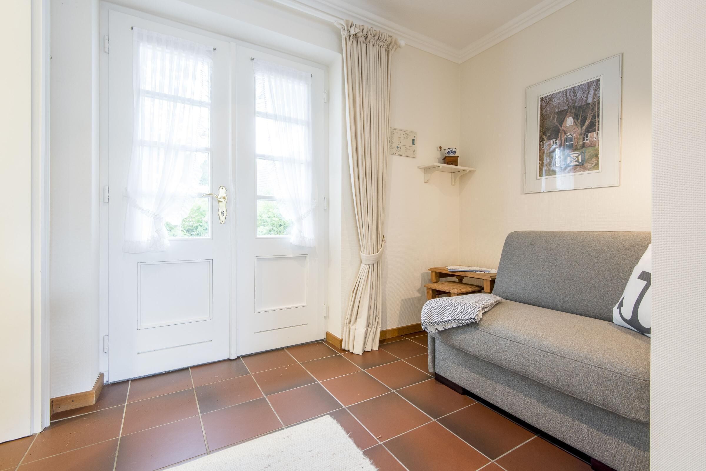 Een kamer met een witte deur, een grijze bank, een klein houten bijzettafeltje en een lijst met een afbeelding aan de muur.