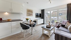 A modern kitchen with white cabinets, a dining table with chairs, a sofa, and a TV. - Stylish. Terrasse. Strandnah. Objekt-id: Hf/03 (Westerland)
