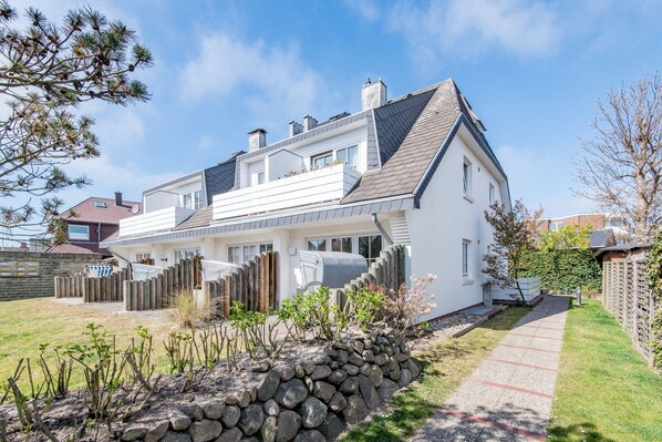 A two-story house with a sloped roof, a stone wall in front, and a pathway leading to the entrance.