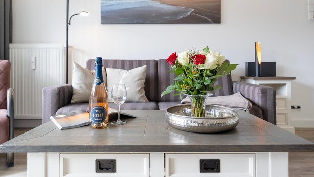 A room with a sofa, a table with a bottle, a glass, and a vase with flowers. - Stylish. Terrasse. Strandnah. Objekt-id: Hf/03 (Westerland)