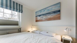 A bedroom with a bed, a nightstand with a lamp, a painting of a beach scene, and a window with a view of trees. - Stylish. Terrasse. Strandnah. Objekt-id: Hf/03 (Westerland)