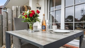 An outdoor dining table with wicker chairs, a vase of flowers, a bottle of champagne, and a glass. - Stylish. Terrasse. Strandnah. Objekt-id: Hf/03 (Westerland)
