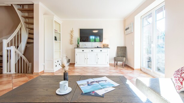 Un vaste espace de vie agrémenté d’une table basse en bois, d’un meuble blanc avec un téléviseur à écran plat, d’un fauteuil en osier et d’une grande fenêtre laissant entrer la lumière naturelle.