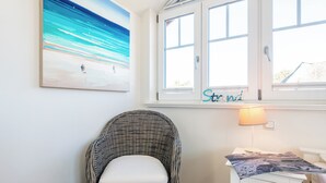 A room with a wicker chair, a nightstand, a lamp, and a painting of a beach scene. - Exklusiv. Garten. Sauna. Objekt-id: Gkw/4b (Westerland)