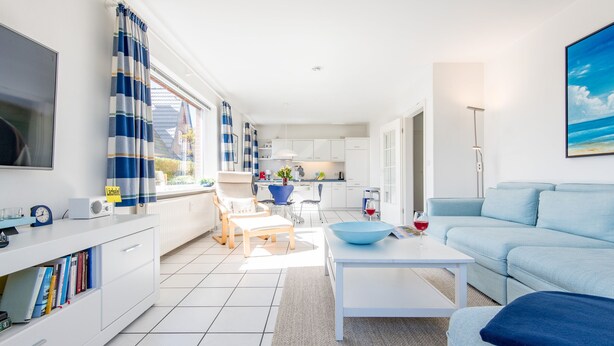 Una sala de estar moderna con sofá, mesa de centro y televisor. Hay una cocina con armarios y una mesa de comedor.