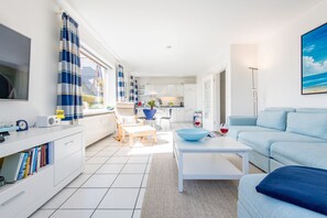 A modern living room with a sofa, coffee table, and a TV. There is a kitchen area with cabinets and a dining table. - Ruhig. Strandnah. Sonnig. Objekt-id: Hks/02 (Westerland)
