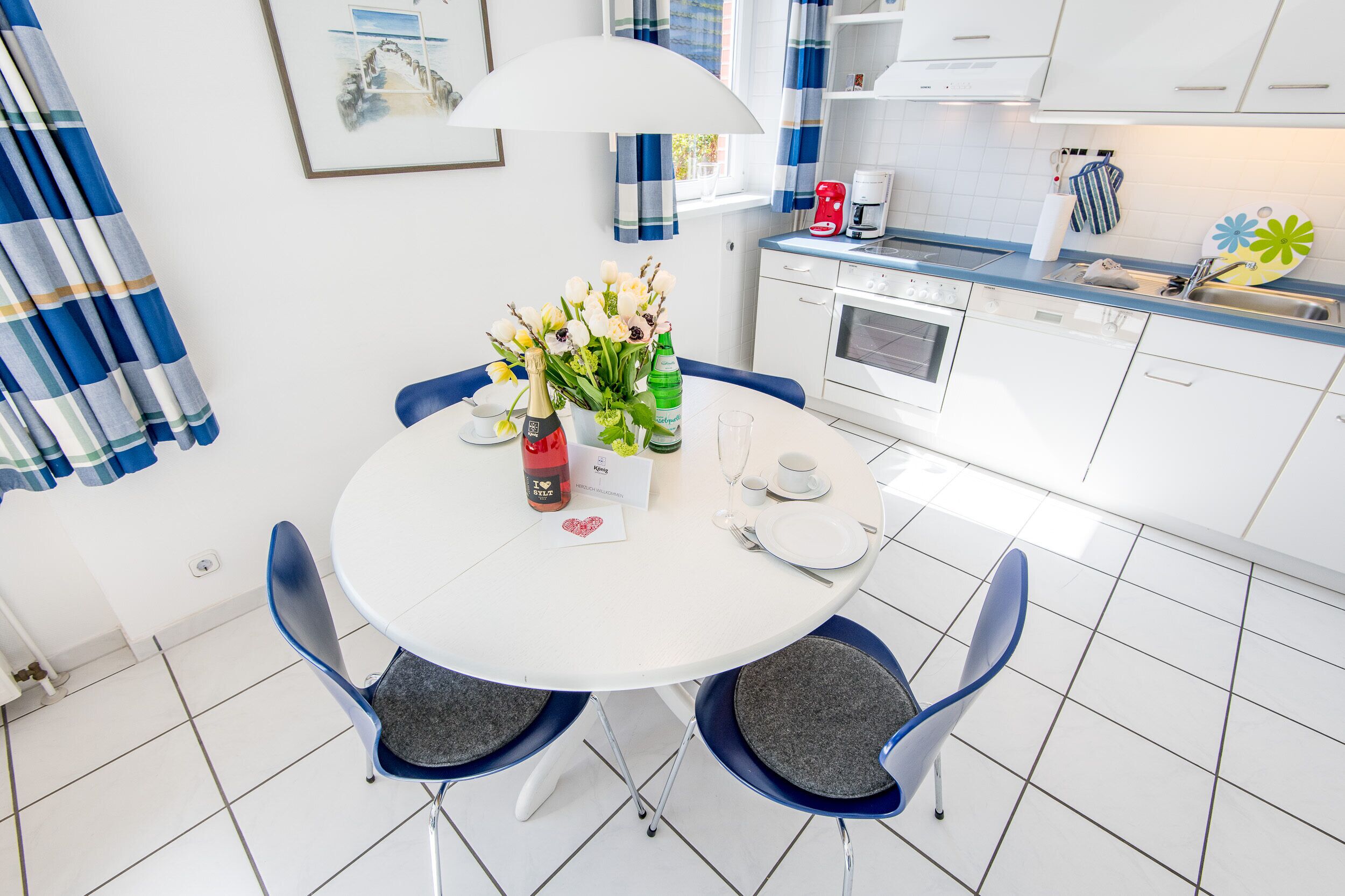 Een moderne keuken met witte kasten, een eettafel met stoelen, een koffiezetapparaat en een ingelijst kunstwerk aan de muur.