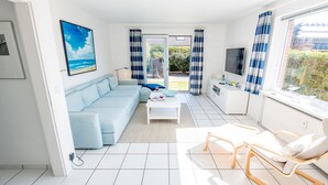Un salón luminoso con un sofá azul claro, una mesa de centro blanca y una silla de madera. Hay un televisor sobre un mueble blanco y un cuadro de una playa en la pared.
