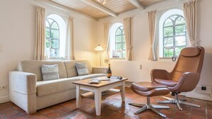 A living room with a beige sofa, a wooden coffee table, a brown leather armchair, and a floor lamp. There are large windows with curtains. - Ruhig. Edel. Garten. Haus Mühlenhof, Whg. 4 Objekt-id: Mht/04 (Westerland)